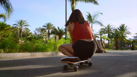 Two-female-friends-on-skateboards-rides-downhill-at-the-resort-having-fun-moving-and-smiling.-Road-along-palm-trees