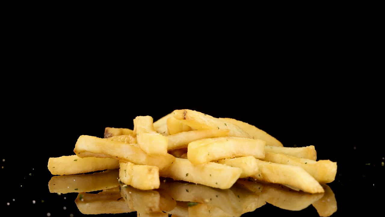 Golden French fries rotate smoothly on a reflective black surface, illuminated by studio lighting. The camera maintains a consistent angle, emphasizing texture and seasoning
