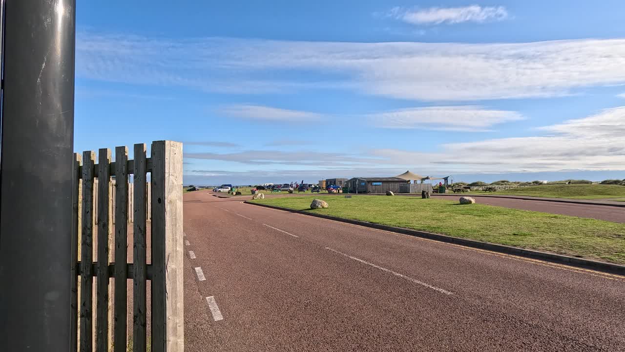 Camera moves past entrance sign toward open road, grassy field, and blue sky, daylight