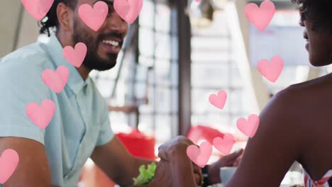 Pink-heart-icons-against-african-american-couple-holding-hands-while-sitting-in-a-restaurant