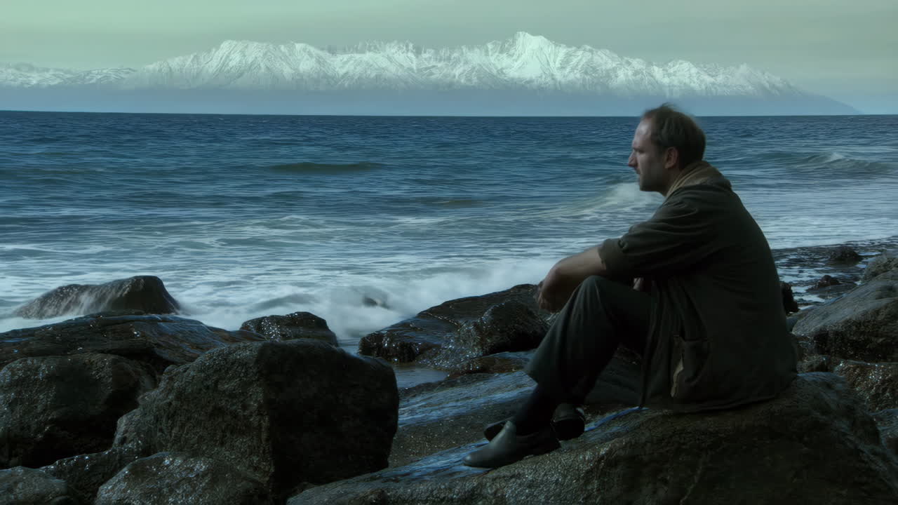 Man Sits on Rocks by the Ocean with Snow-Capped Mountains in the Distance