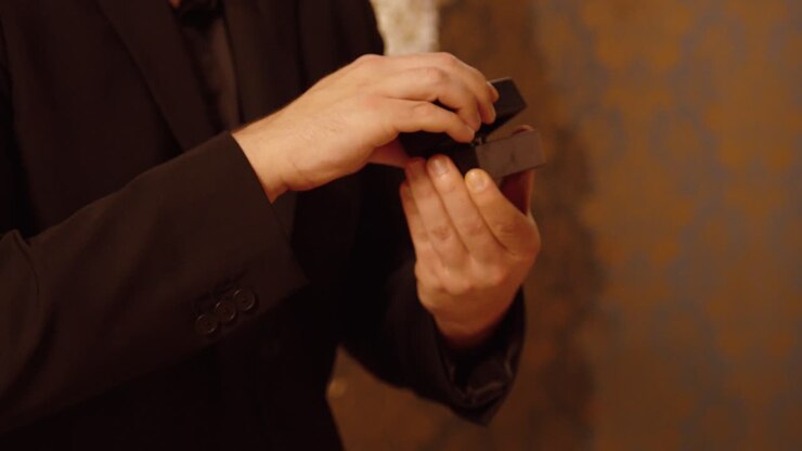 Un uomo in abito nero estrae delicatamente un piccolo portagioie dalla tasca della giacca, preparandosi per una romantica proposta in un'elegante atmosfera illuminata