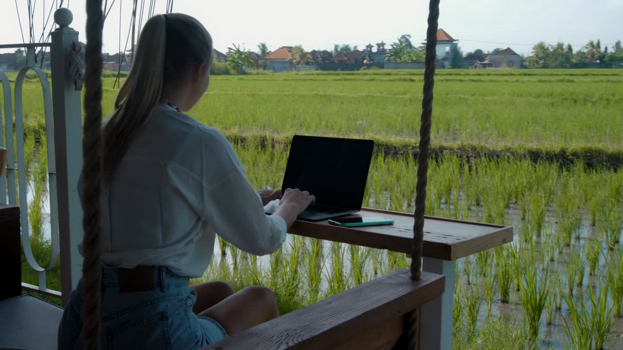 青いドレスを着た若いヨーロッパの女の子がラップトップでオンラインでリモートで作業し緑の植物の裏庭のスクリーンを見ています