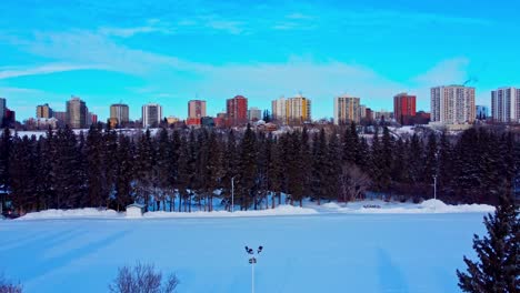 early-morning-aerial-rise-winter-handful-of-ice-skaters-at-Canada's-largest-manmade-skating-rink-surrounded-by-tall-trees-with-residential-condos-apts-overlooking-Victoria-Park-in-Edmonton-Alberta