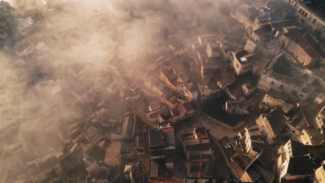 drone shot of old Italian city town - Matera. The city is shrouded with low handing clouds, buildings are visible through them. Warm colours of the rising sun reflected against buildings