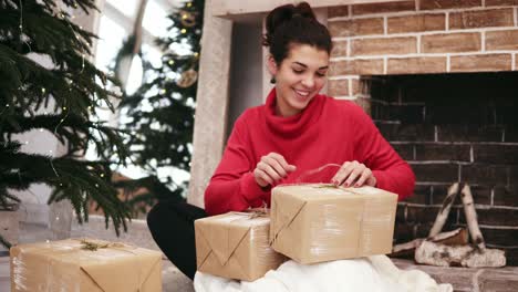 Hermosa-Mujer-Sonriente-Envolviendo-Regalos-De-Navidad-Sentada-Junto-Al-árbol-De-Navidad-Y-La-Chimenea-En-Casa-Y-Luego-Poniéndolos-Debajo