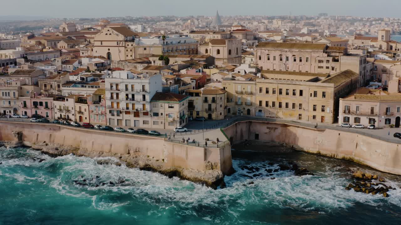 Aerial drone shot of Ortigia island in Sicily. Dolly out drone shot with large waves crashing on the rocks. Tilt up drone shot revealing the city of Syracuse. UNESCO world heritage site in Italy.