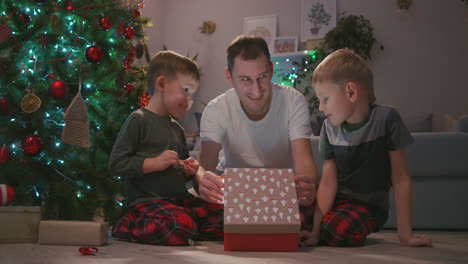 father with two children opens a christmas present under the tree and rejoices. a gift for my father. a ray of light from the gift shines on the face. rejoice with the children new year's gift. opening a gift together. high quality 4k footage