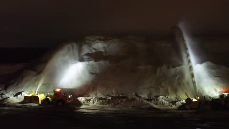 Pair-of-blowers-suck-snow-up-and-shoot-it-out-into-high-tall-pile-at-night,-flood-light-illuminating-area