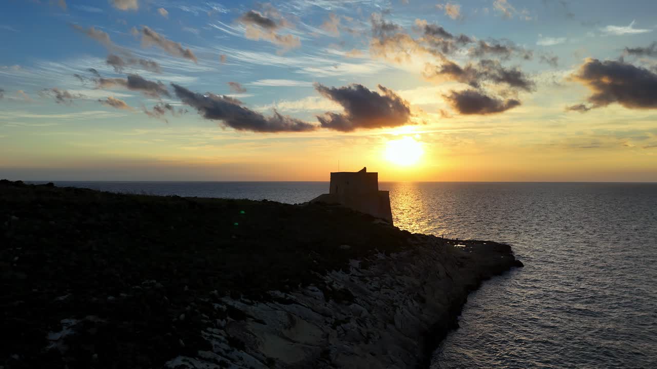 Silhouette drammatica al tramonto della storica torre di Qolla l-Bajda su una scogliera frastagliata di Gozo, con la luce dorata del sole che si riflette sul Mar Mediterraneo sotto un cielo nuvoloso e sparso a Malta