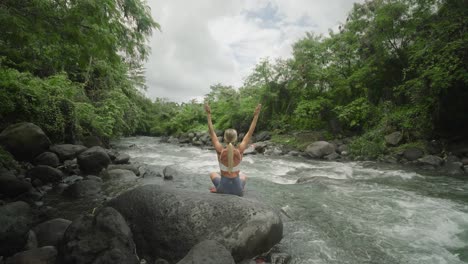 behind blond fit woman raising arms while in easy pose on rock beside tropical river