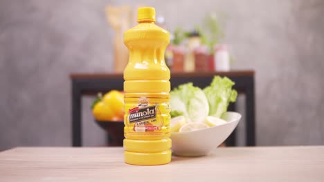 hand gets minola lauric oil in a yellow plastic bottle from the wooden table with a bowl of fresh fruits and vegetables - closeup shot