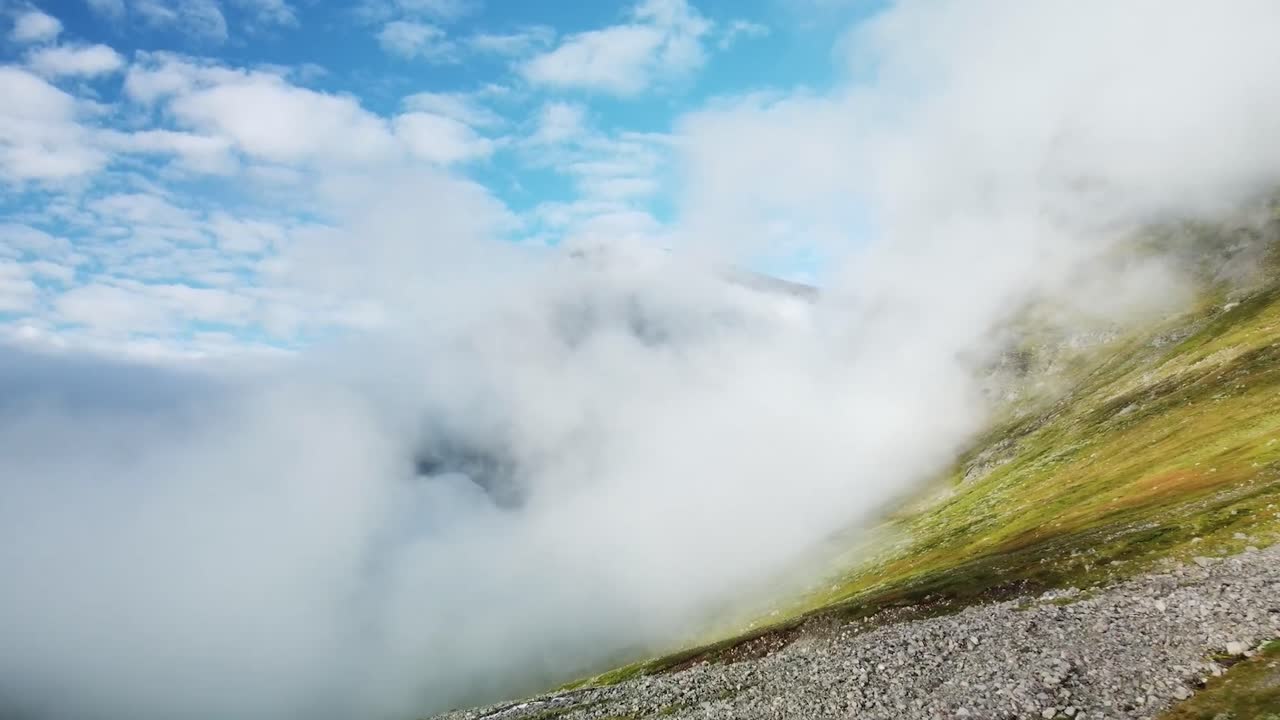 Aerial low altitude footage flying above green and brown colored hillside or mountain rocky terrain during a sunny day in Sweden where white fluffy clouds are low above the ground. Blue sky visible.