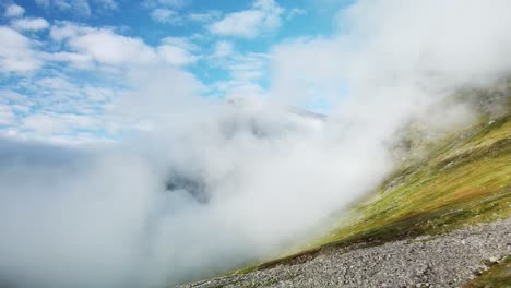 aerial low altitude footage flying above green and brown colored hillside or mountain rocky terrain during a sunny day in sweden where white fluffy clouds are low above the ground. blue sky visible.