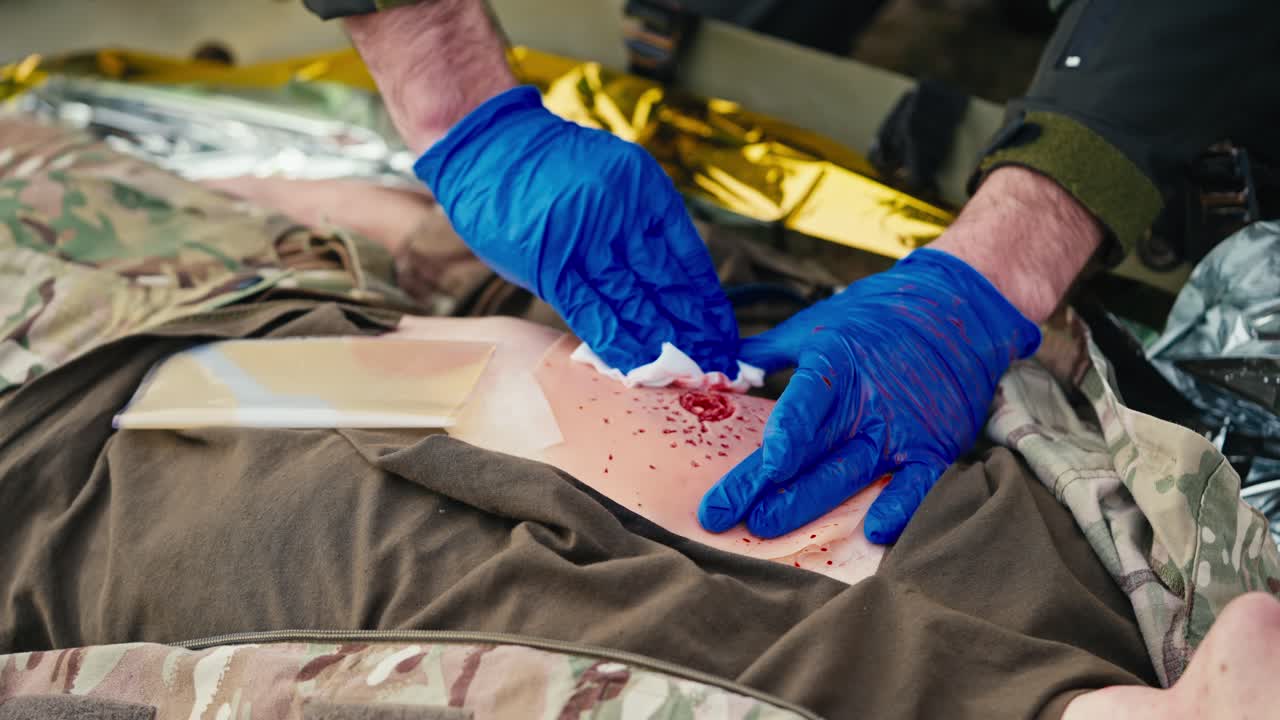 자신감 넘치는 군의 의사, 손에는 파란색 고무 장갑을 착용하여 부상당한 병사의 가을 똑바로 펴고 특별한 플래스터를 사용하여 출혈을 멈추고 전투에서 응급 처치를 제공합니다.