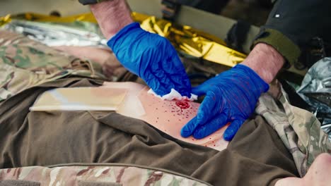 un médico militar confiado con guantes médicos de goma azul en sus manos endereza el pecho de un soldado herido y detiene la hemorragia usando un yeso especial mientras proporciona primeros auxilios en combate