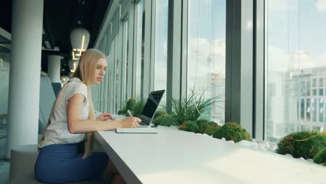 formal woman with laptop and papers. side view of businesswoman writing on paper while sitting with laptop at table near window in modern office