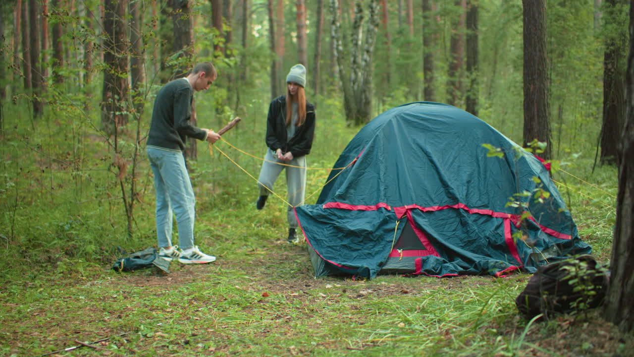 Young man in sweater tries to separate tangled tent ropes while woman joins to assist him during forest camping session, both coordinating effort to stabilize setup in lush green outdoor woodland area