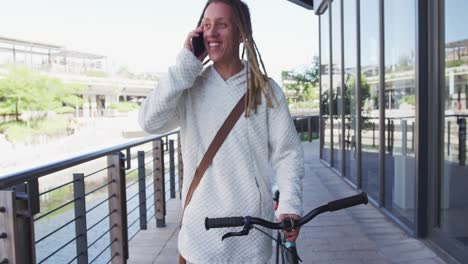 Mixed-race-man-with-dreadlocks-wheeling-a-bicycle-in-the-street-talking-on-smartphone