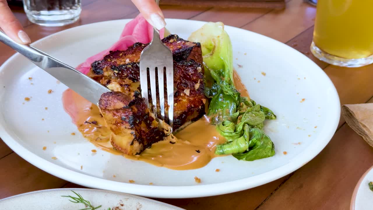 A hand uses a fork and knife to slice into a glazed pork belly steak served with sauce and greens on a white plate under bright lighting
