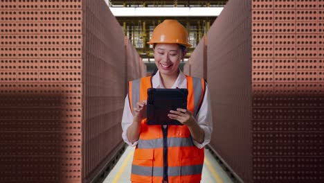 asian female engineer with safety helmet working on a tablet while standing with red brick packed in stacks are stored