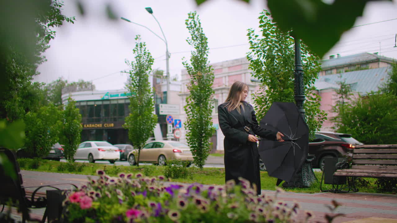 Gorgeous woman in long black coat struggles with umbrella on windy rainy city street near bench and flowers, looking down as stormy weather surrounds her with cars passing in urban background atmosphere