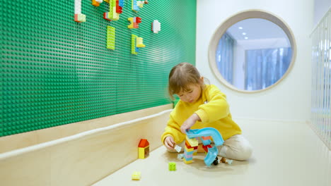 Kleines,-Süßes,-Kaukasisches-Amerikanisches-Mädchen,-Das-Mit-Bunten-Legosteinen-Spielt-Und-Eine-Rampe-Baut,-Die-Im-Indoor-Spielzimmer-Auf-Einem-Boden-Sitzt---Zeitlupe,-Breit