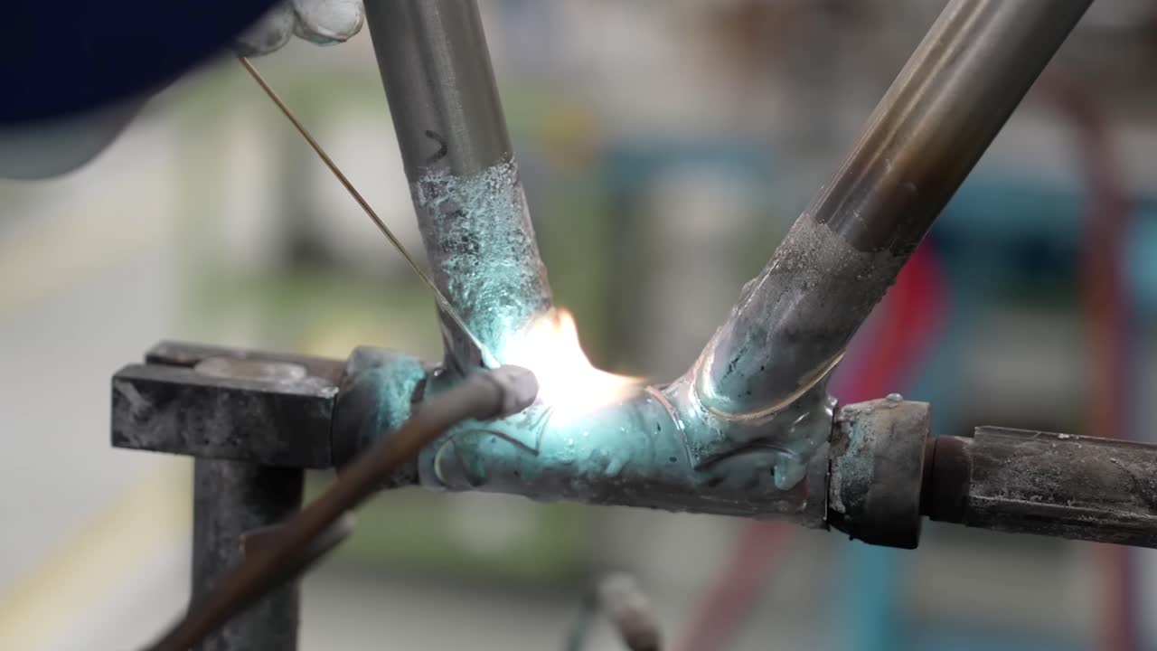Primo piano di un artigiano professionista che brasa manualmente un giunto di un telaio di bicicletta con raccordi, usando una fiamma ossidrica e una bacchetta d'apporto.