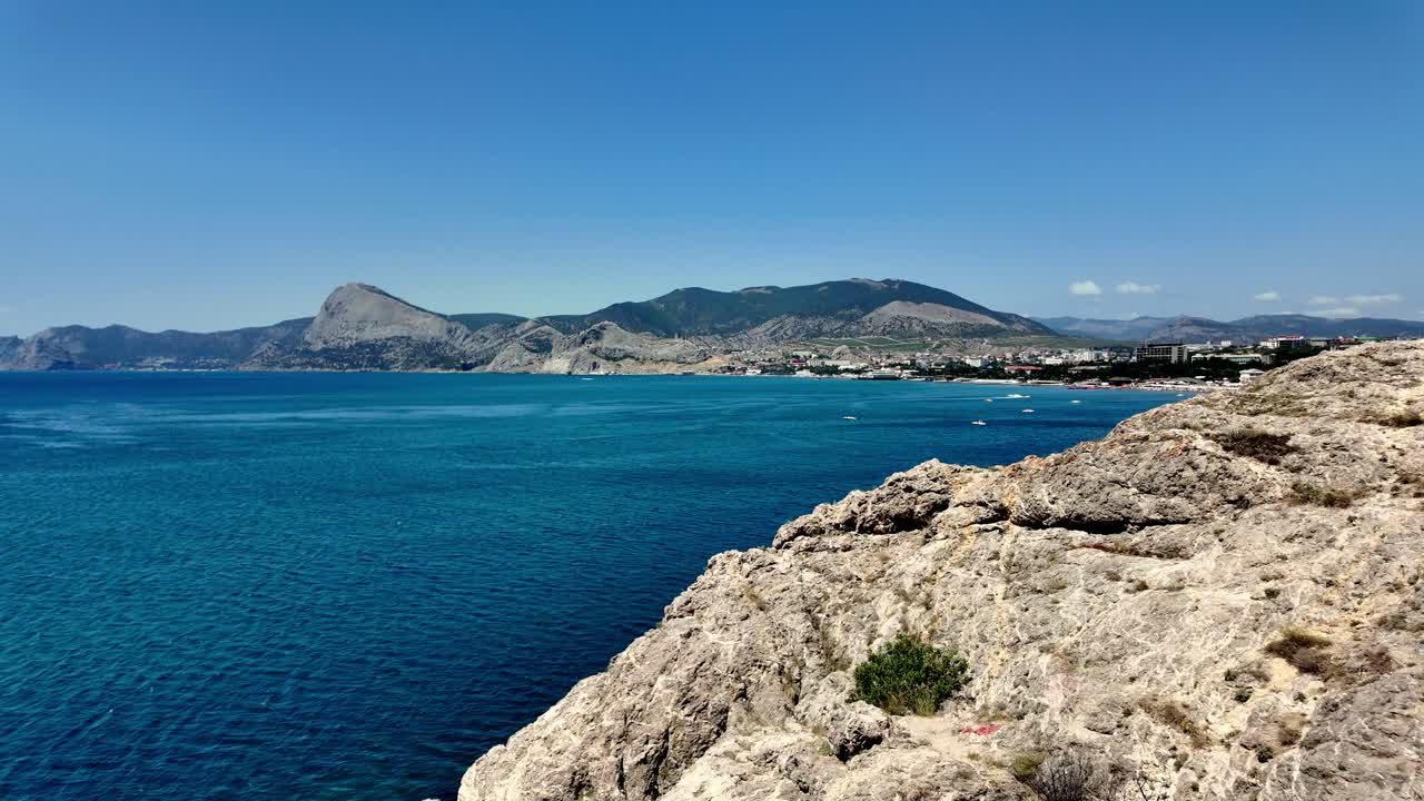 A panoramic view of Sudak, Crimea, Russia, featuring a clear blue sky, the Black Sea, and a rocky coastline