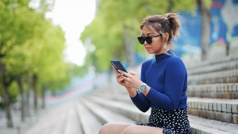 Woman-in-a-blue-sweater-and-sunglasses-takes-a-selfie-while-sitting-on-outdoor-steps