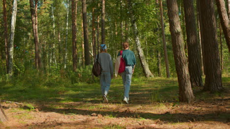 vista trasera de dos amigos caminando en un bosque verde exuberante llevando mochilas, uno con corbata de cabello azul y otro con tela colgada en la bolsa, rodeados de árboles altos y luz solar manchada