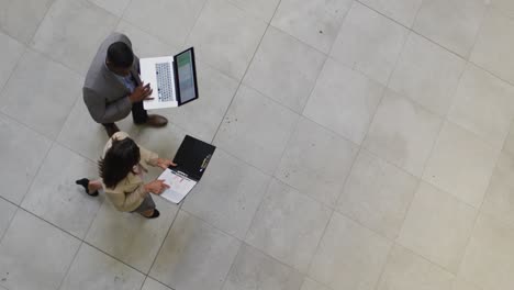 Diverse-businessman-and-businesswoman-walking-with-laptop-in-lobby-of-modern-office