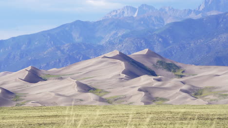 Stunning-late-summer-telephoto-view-of-The-Great-Sand-Dunes-National-Park-Colorado-Rockies-mountain-sandy-14er-peaks-crisp-golden-yellow-tall-grass-wind-clouds-blue-sky-mid-day-cinematic-down-pull