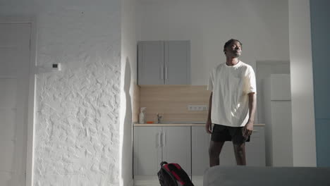 man in modern kitchen holding black backpack, preparing to leave, sunlight casting shadows on white walls, simple interior with kettle, sink, and kitchen cabinets in background