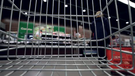 shopping trolley is moving quickly along aisles in supermarket, shelves with food, drinks and goods are visible through cart grille. shopping and customers concept.