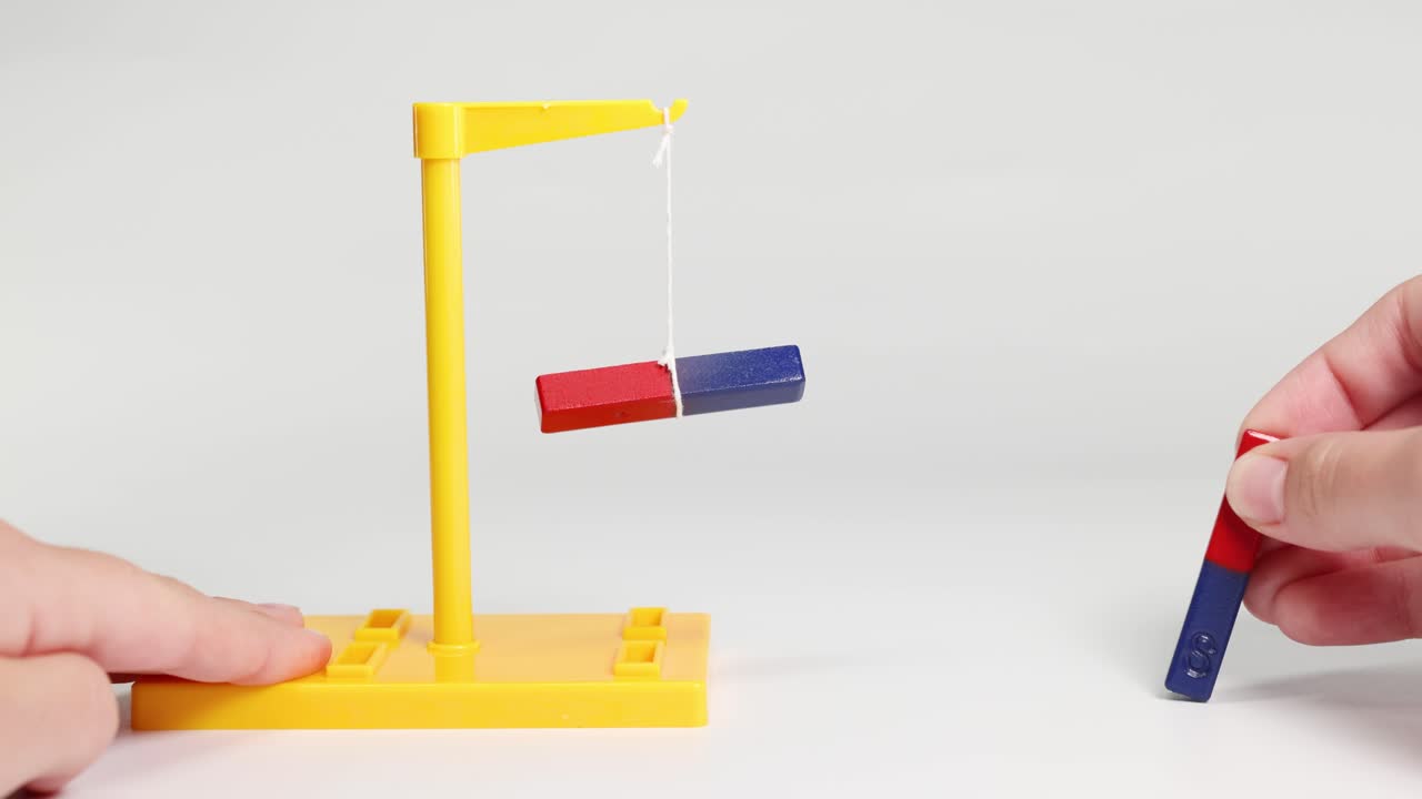 Hands manipulate magnets to demonstrate attraction and repulsion on a yellow stand. Neutral lighting highlights the magnetic forces