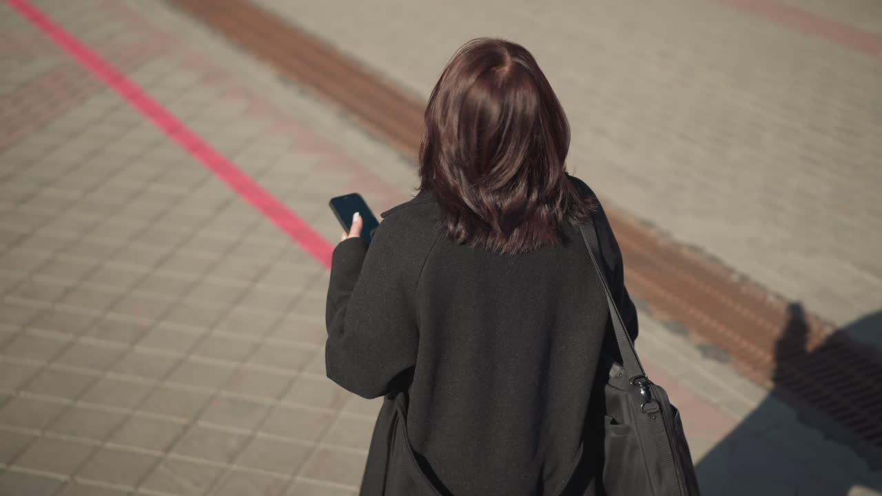 전화기에 집중하면서 머리카락을 만지고 바닥에 그늘을 던져 주위를 둘러보는 여성의 뒷면.