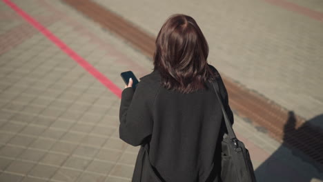 back view of a lady walking outdoors on an interlocked path, touching her hair while focused on her phone and looking around with a shadow cast on the ground