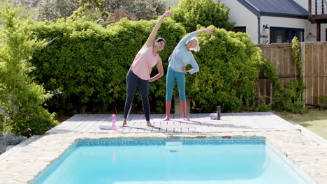 two happy diverse senior women stretching and smiling by pool in sunny garden, slow motion