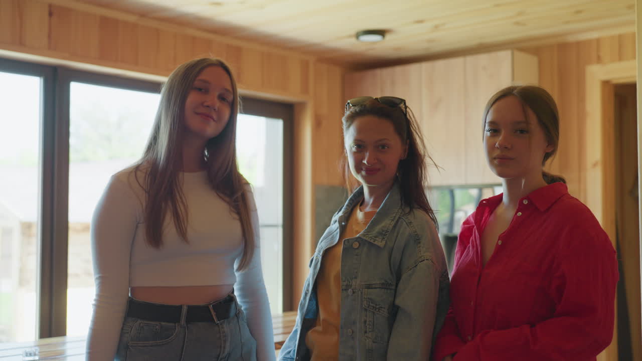 Three stylish women pose together inside wooden cabin as middle one lifts sunglasses off her head while others smile gently in relaxed setting with bright natural light pouring through glass windows