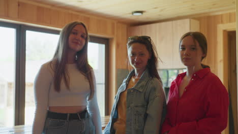 three stylish women pose together inside wooden cabin as middle one lifts sunglasses off her head while others smile gently in relaxed setting with bright natural light pouring through glass windows