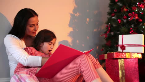 mother reading her daughter a story by the christmas tree