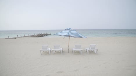 Paraguas-Azul-Y-Blanco-En-Una-Playa-Solitaria-Y-Ventosa-Con-Arena-Blanca-Tropical-Y-Cielo-Gris,-Establecida