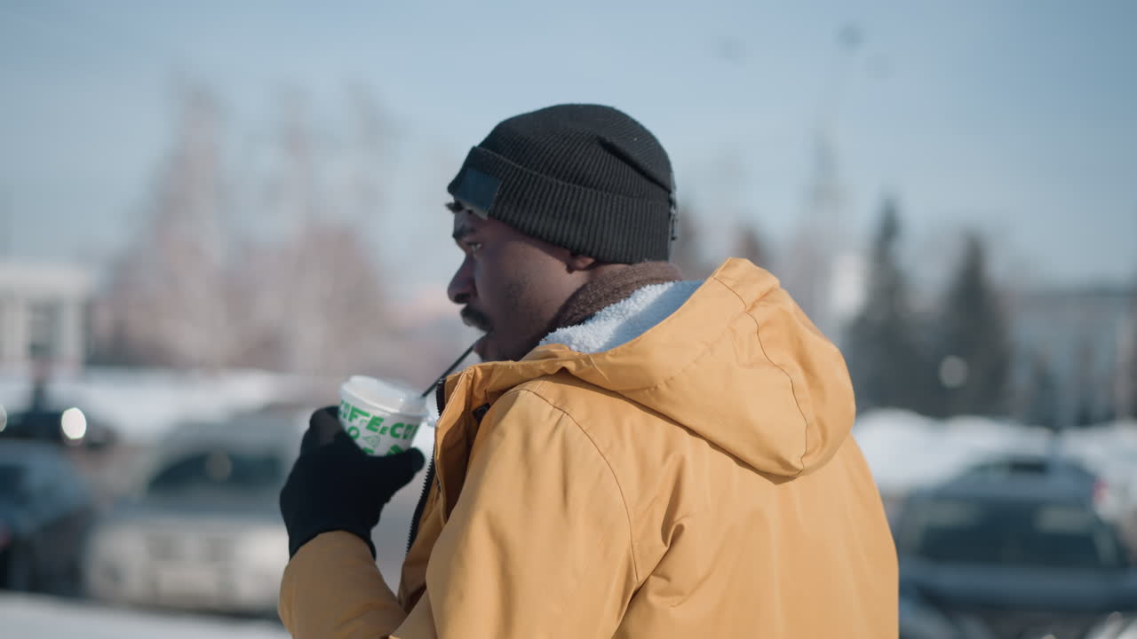 high aerial view of photographer sipping coffee wearing gloves and jacket in winter cold, holding camera while scanning urban snowy streets under morning sun, visible breath rising in crisp air