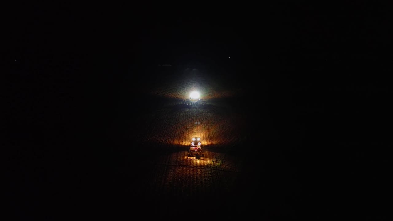 Trattore esegue la semina notturna utilizzando fari luminosi che illuminano le file di colture attraverso un campo agricolo buio a Formosa, Goiás, Brasile, mostrando agricoltura di precisione e funzionamento continuo, ripresa aerea