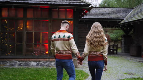Beautiful young couple walking along wooden building in the rural district. Love story, romantic atmosphere. Two lovers holding hands in the same sweaters.
