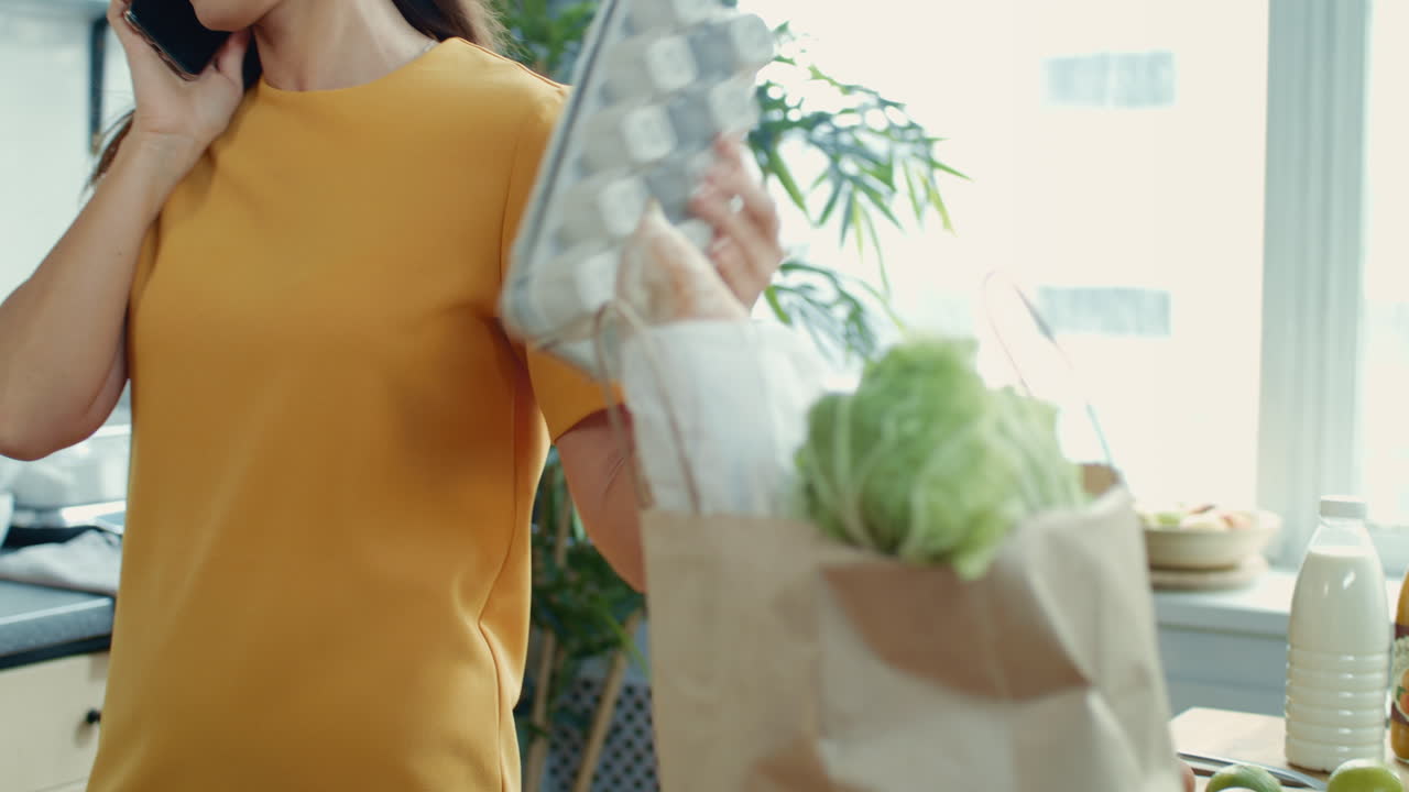 una mujer enfocada hablando por teléfono en la cocina. una chica desempaquetando una bolsa de compras en casa.