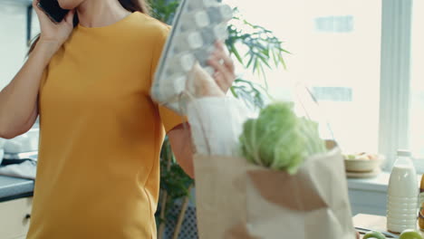 focused woman talking phone in kitchen. girl unpacking shopping bag at home.