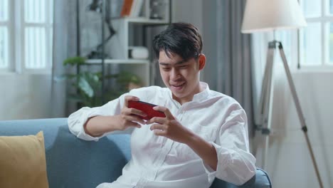 Asian Man Playing Game On Smartphone And Smiling While Lying On Sofa In The Living Room