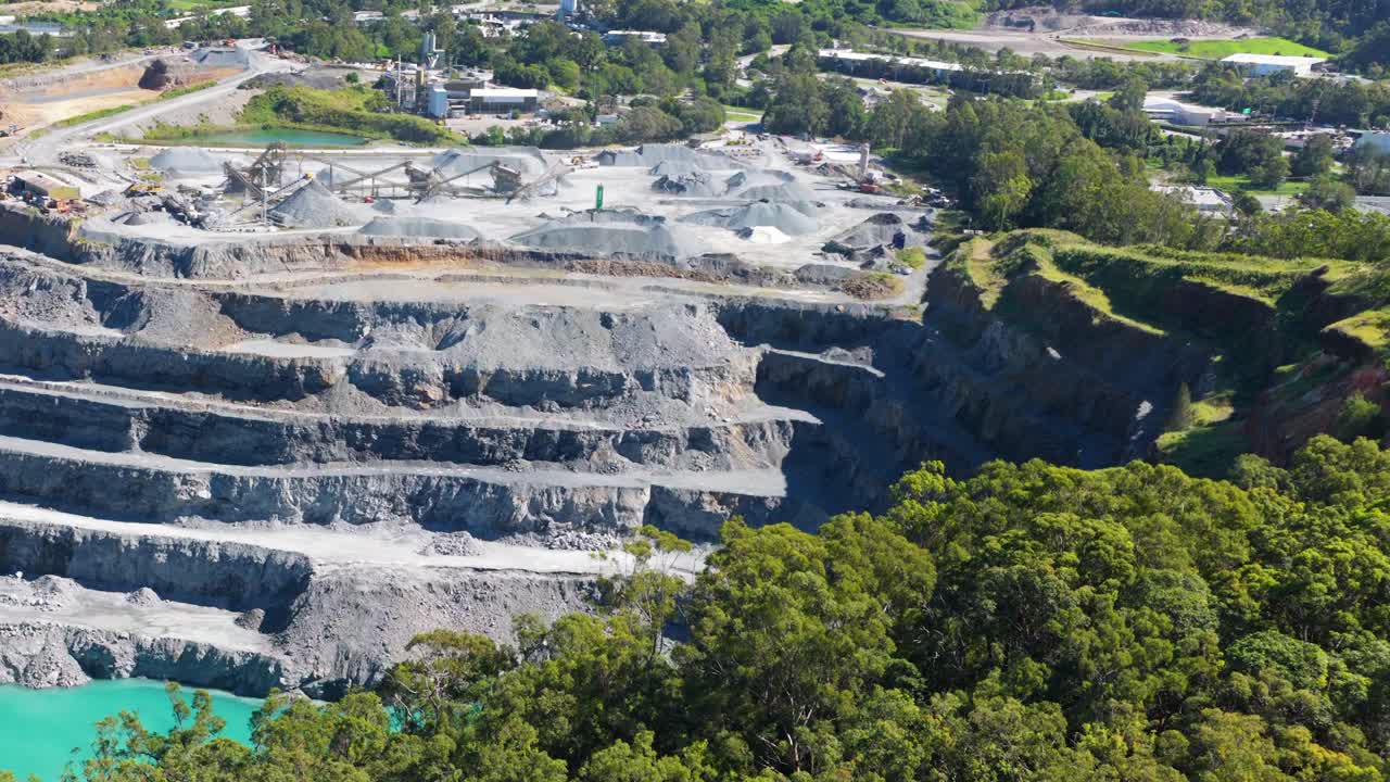 Aerial footage captures a large quarry surrounded by dense greenery under bright daylight, highlighting industrial and natural contrasts